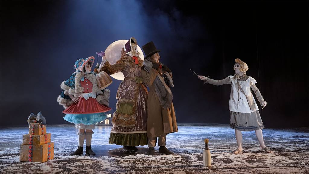 On a snowy winter night, three dancers, the Donnarumma family stand together looking to the right at the little match girl who is wearing a white bonnet and pinafore and is holding out a match.